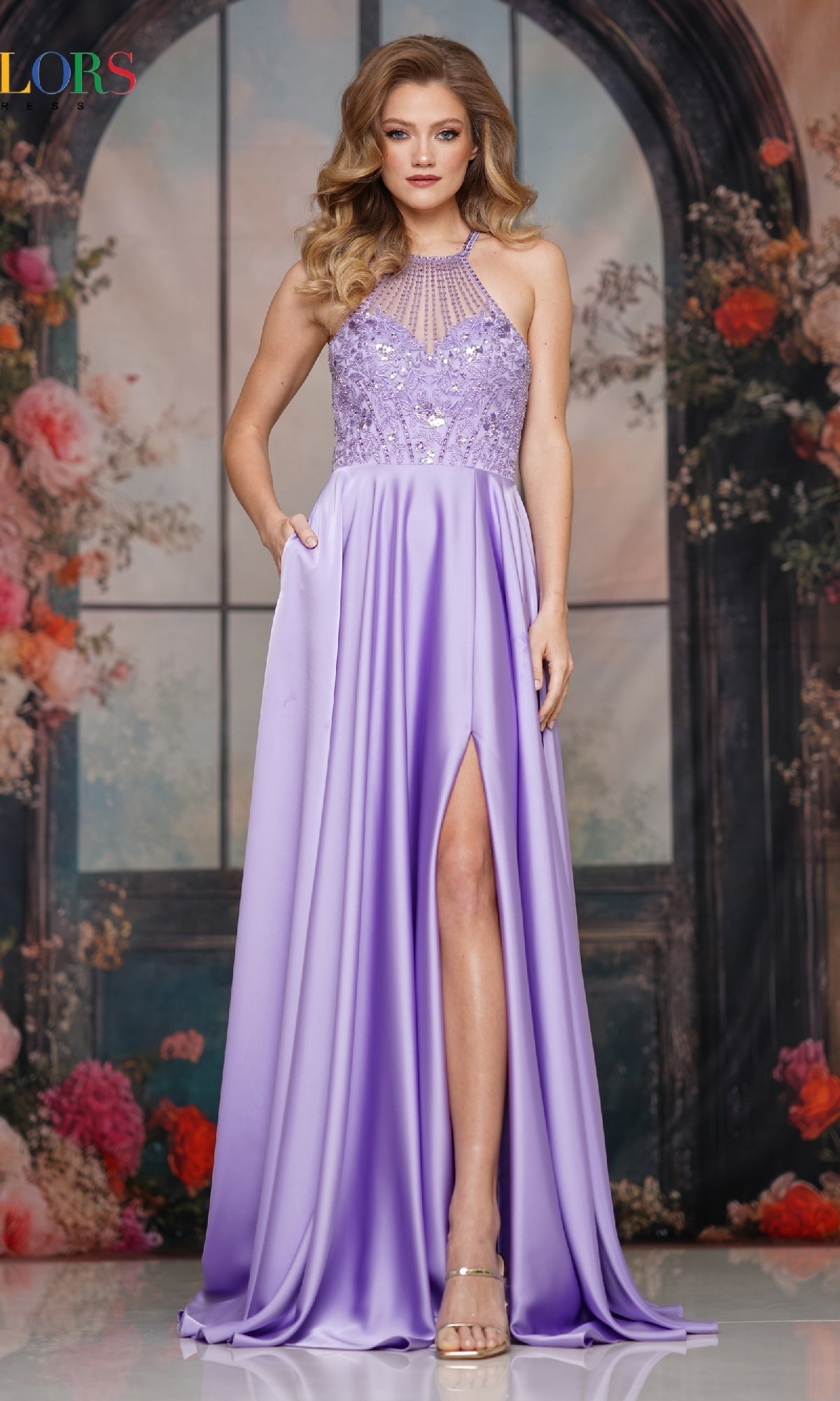 A woman in the Colors Dress High-Neck Halter Prom Gown (Style 3822) with sequin beading and a high slit stands before a floral arch. She pairs the lavender dress with high heels and wears her hair in loose waves over her shoulders.